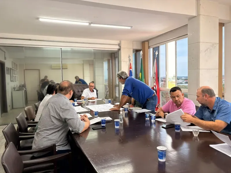 Adultos participam de reunião ao redor de uma mesa com documentos. A imagem acompanha comunicado municipal sobre o plano de recuperação da malha asfáltica.